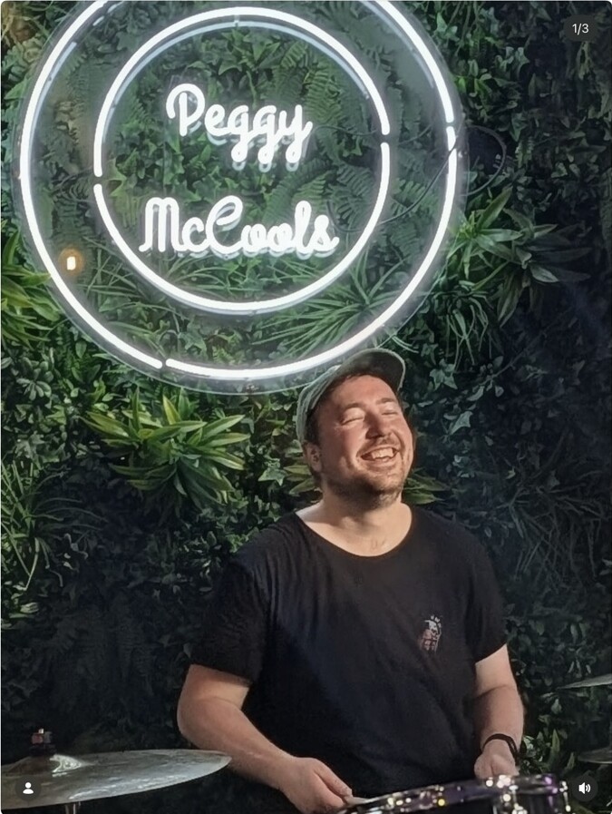 A smiling drummer performing beneath the Peggy McCools sign